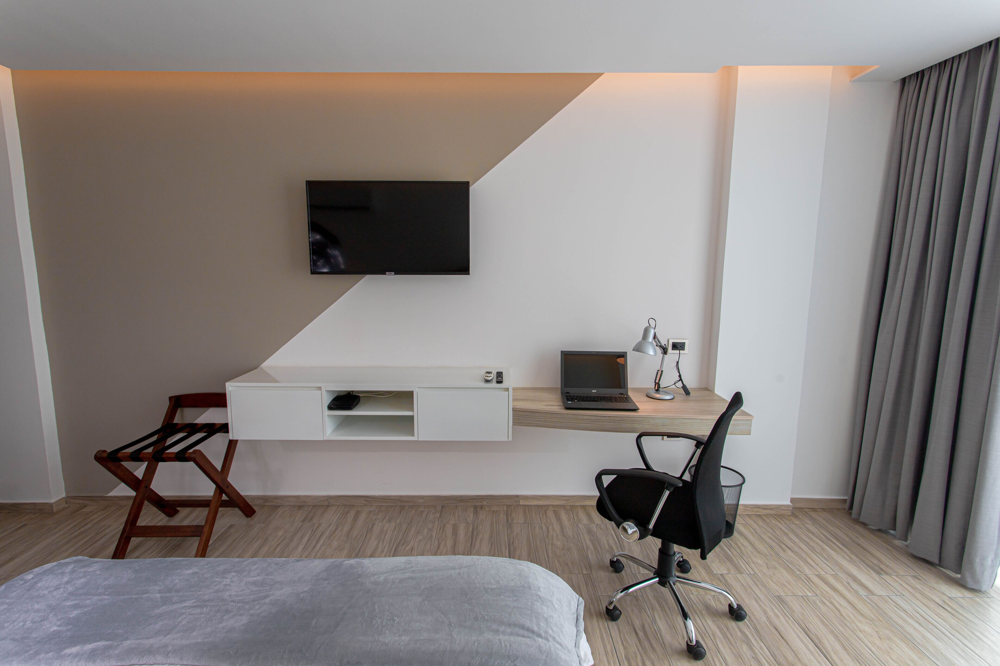 A hotel room with a bed, a desk with a laptop, a chair, a TV mounted on the wall, and a window with curtains.