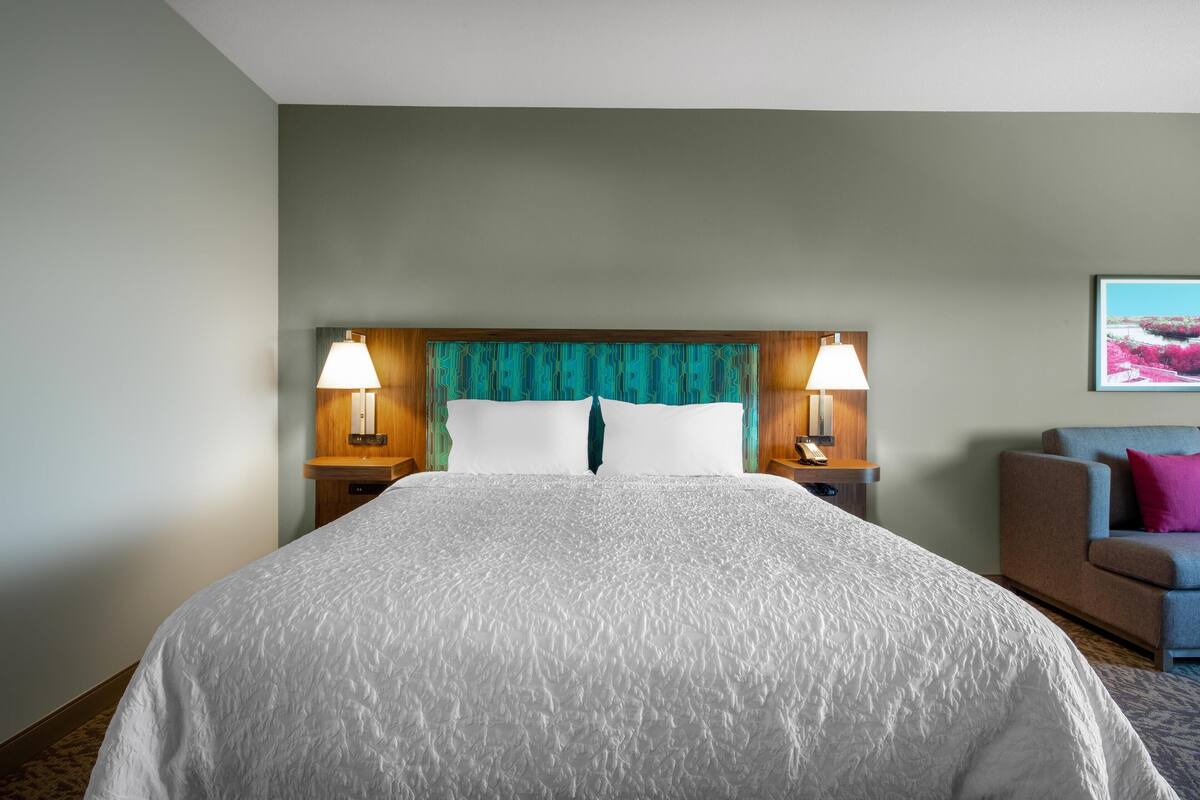 A hotel room with a large bed, two bedside lamps, a wooden headboard, and a chair with a cushion.