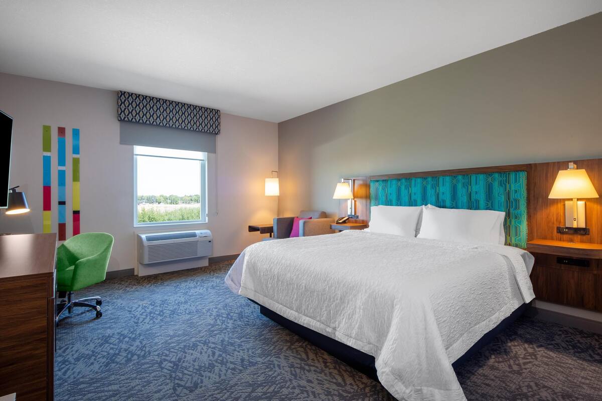 A hotel room with a large bed, a desk with a chair, a window with a view, and a wall with colorful vertical stripes.