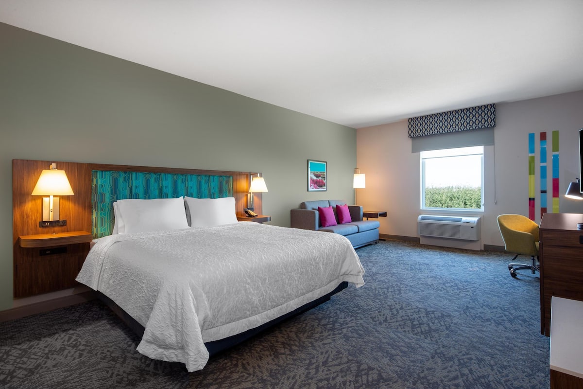 A hotel room with a large bed, two bedside lamps, a sofa, a desk, and a window with a view.
