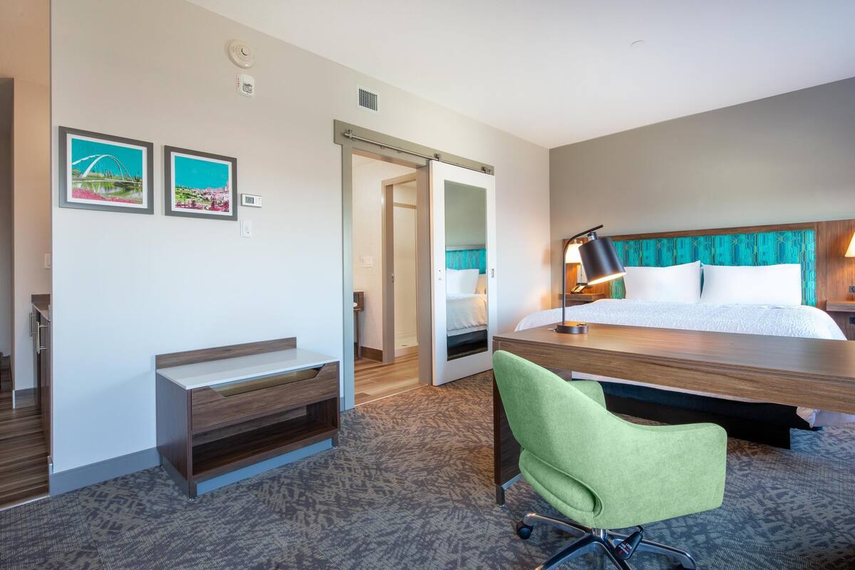 A hotel room with a bed, desk, chair, and two framed pictures on the wall.