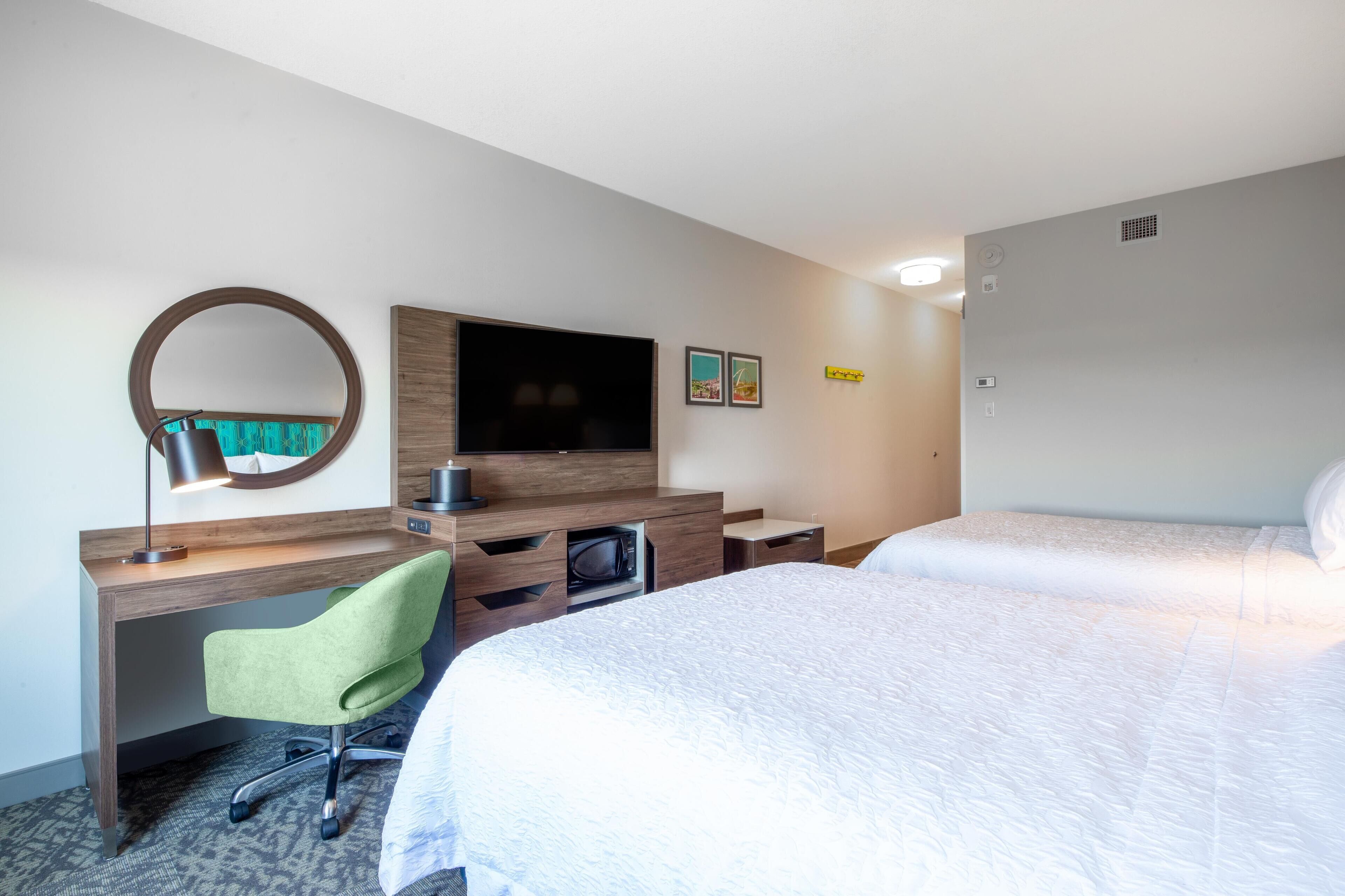 Quarto de hotel com cama, escrivaninha, cadeira, televisão e espelho.