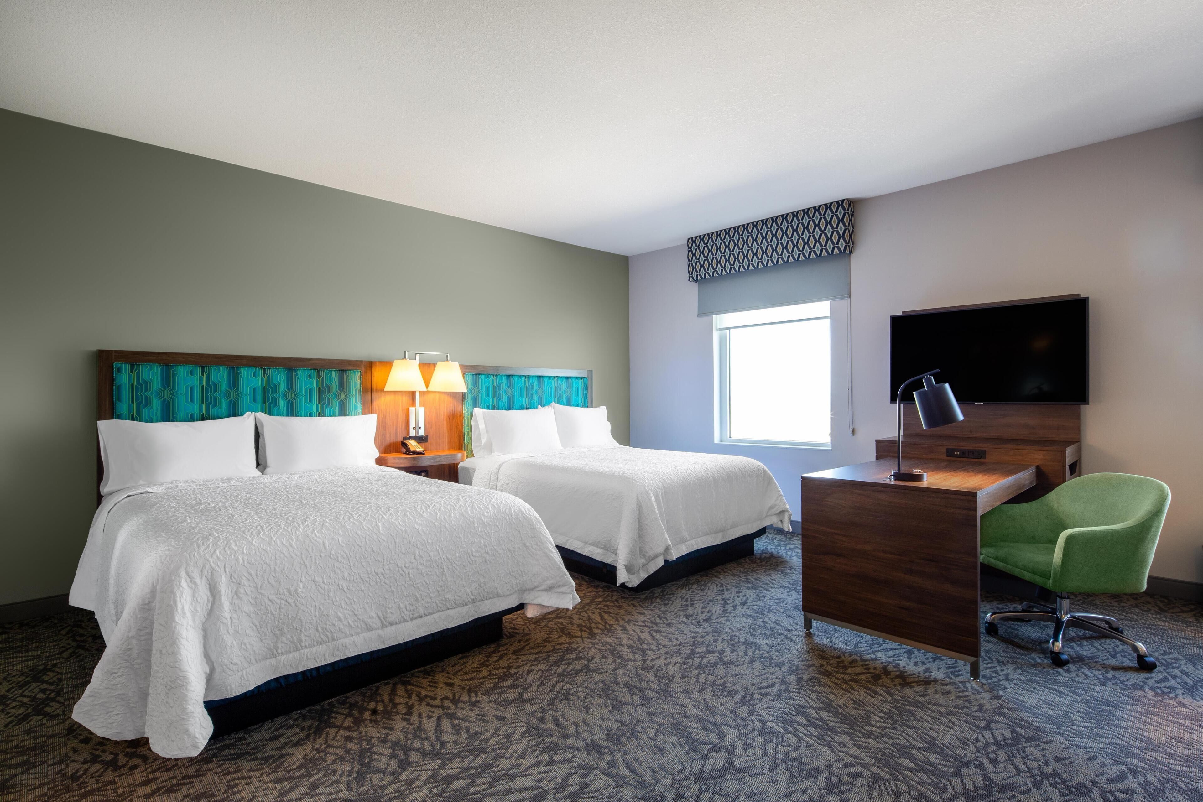 A hotel room with two beds, a desk, a chair, a TV, and a window.