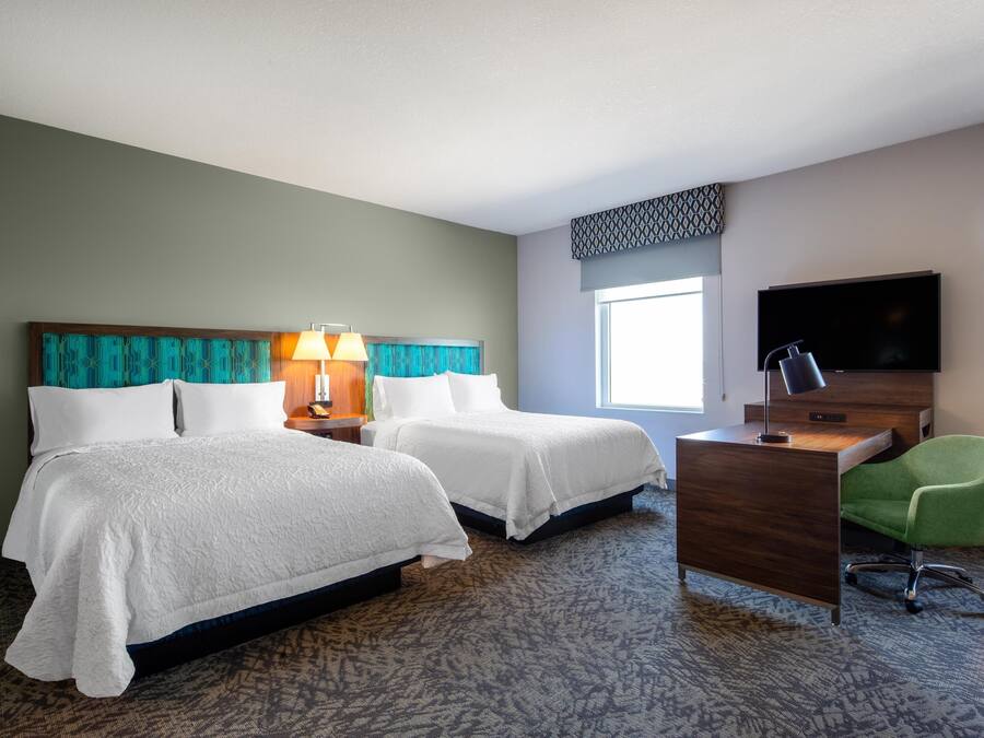 A hotel room with two beds, a desk, a chair, a TV, and a window.