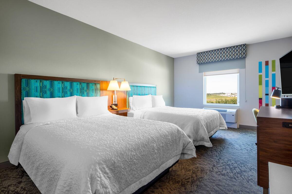 A hotel room with two beds, a desk, a chair, a TV, and a window with a view.