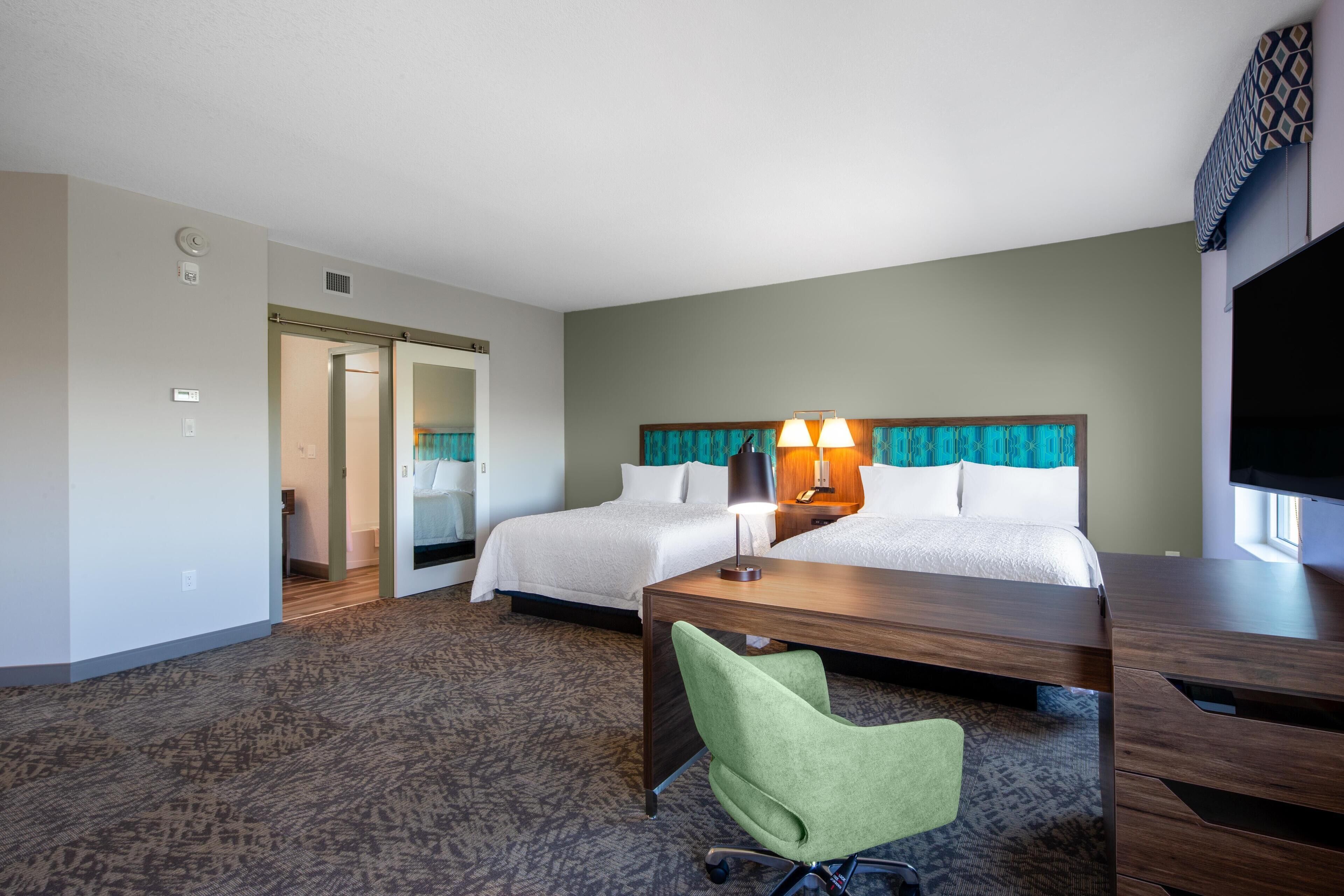 A hotel room with two beds, a desk, a green chair, a TV, and a bathroom with a shower and toilet.