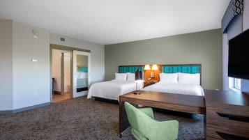 Quarto de hotel com duas camas, uma escrivaninha, uma cadeira verde, uma televisão e banheiro com chuveiro e vaso sanitário.