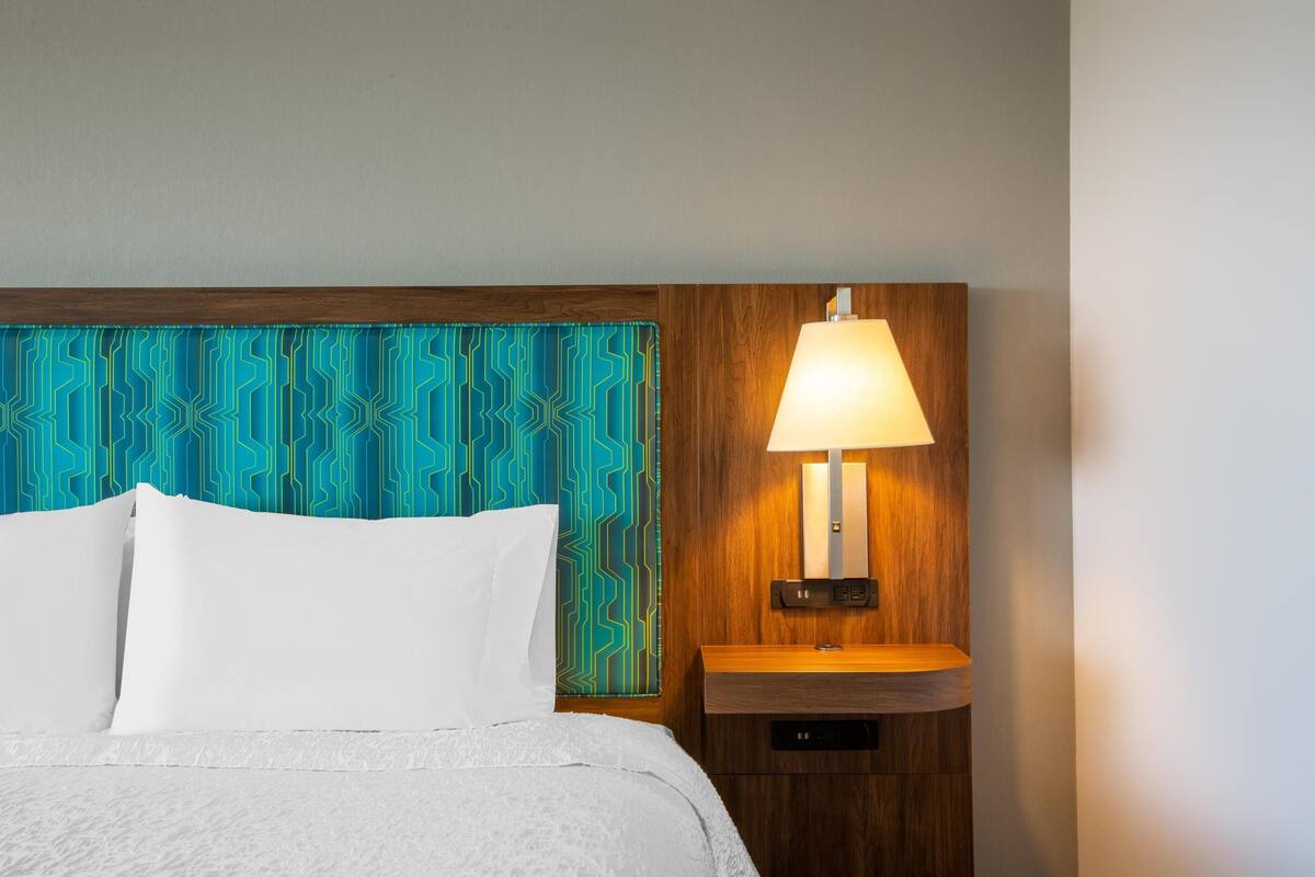 A neatly made bed with white linens, a wooden headboard with a blue geometric pattern, and a bedside table with a lamp.
