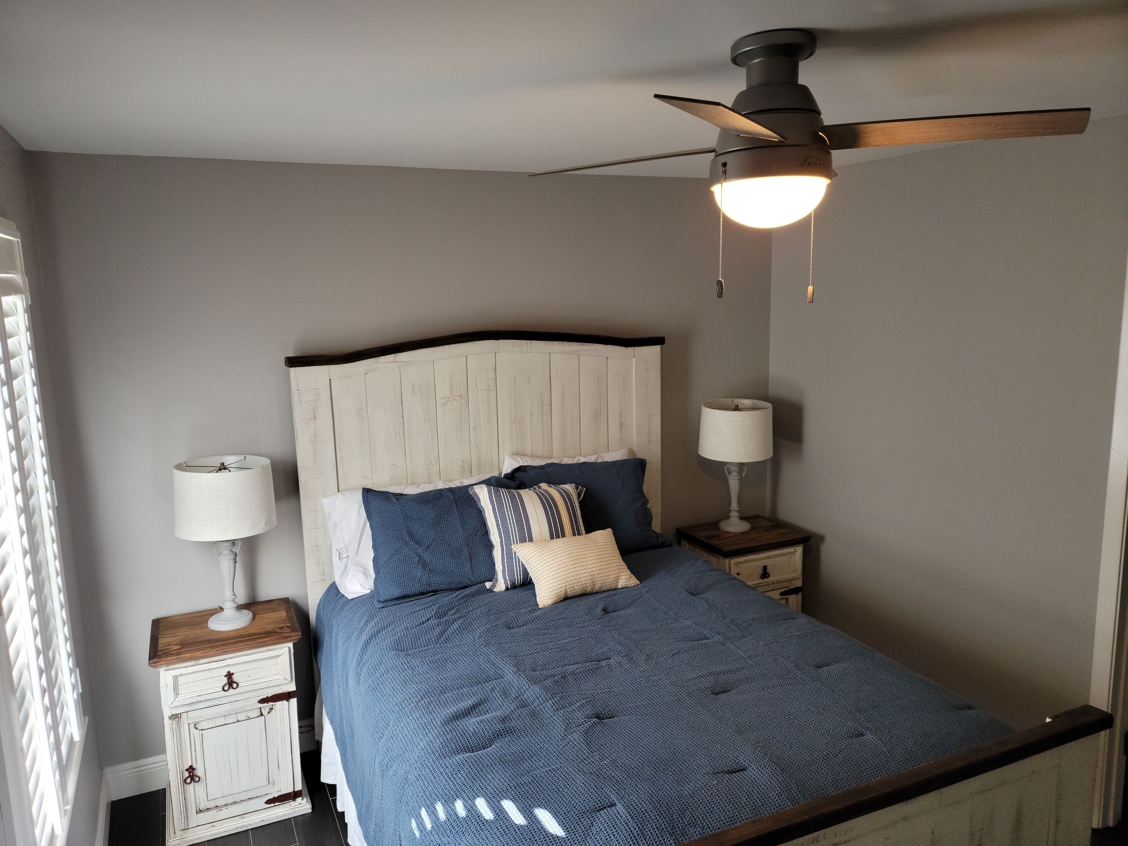 Une chambre à coucher comprenant un lit, deux tables de chevet, un ventilateur de plafond et une fenêtre avec des stores.
