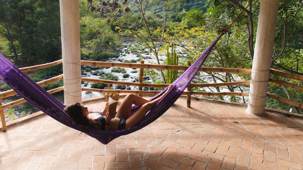 Uma pessoa relaxando em uma rede em um pátio de tijolos, com vista para um rio e vegetação.