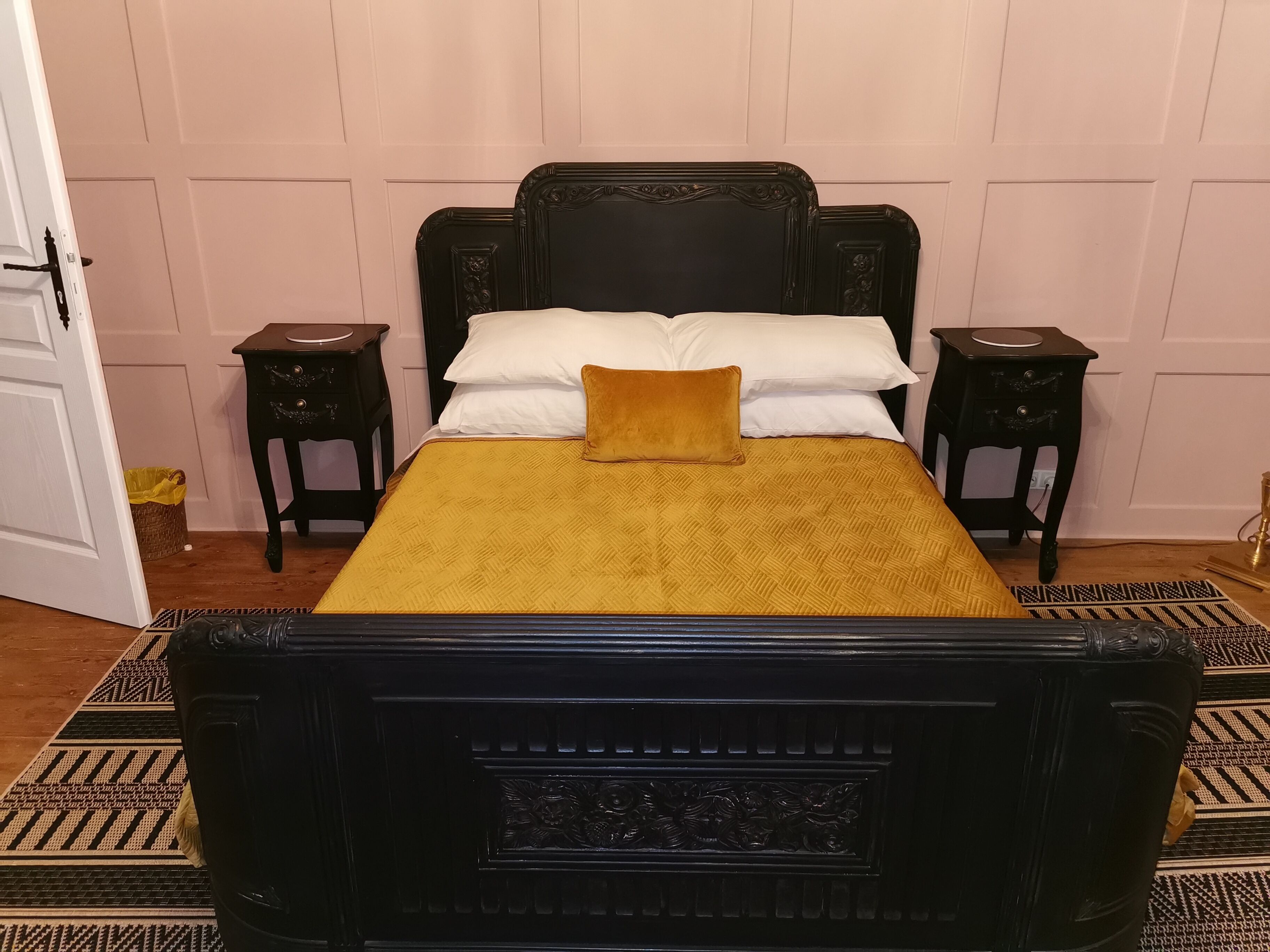 A bed with a black headboard and a mustard-colored bedspread, flanked by two nightstands with drawers, in a room with wooden flooring and a patterned rug.