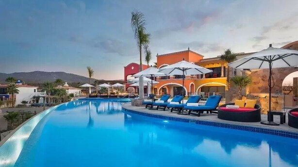 A swimming pool with lounge chairs and umbrellas, surrounded by buildings with arched windows and a mountainous backdrop.
