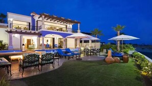 A modern two-story house with a patio area featuring outdoor furniture and umbrellas, a swimming pool, and a well-maintained lawn.