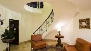 A living room with a curved staircase, two sofas, a patterned rug, a small table with a lamp, and a decorative mirror.