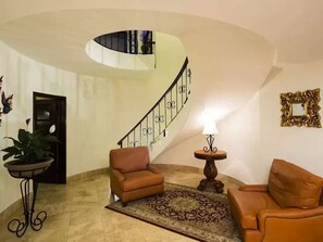A living room with a curved staircase, two sofas, a patterned rug, a small table with a lamp, and a decorative mirror.