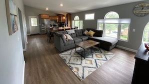 A spacious living room with a gray sectional sofa, a coffee table, and a patterned rug. A kitchen with wooden cabinets and stainless steel appliances is visible in the background. - Sunny Beach Getaway-Outdoor Shower, Big Deck, Walk to Coffee+Ice Cream-Sleeps 10 (Narragansett)