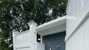 A small outdoor shower area with a white wooden enclosure, a dark shingled wall, and a blue hose. - Sunny Beach Getaway-Outdoor Shower, Big Deck, Walk to Coffee+Ice Cream-Sleeps 10 (Narragansett)