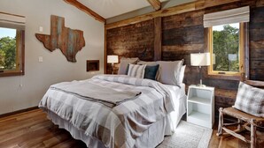 Ein Schlafzimmer mit Holzbalkendecke, einem großen Bett, einem Nachttisch, einem Sessel und einem Fenster mit Blick auf Bäume.