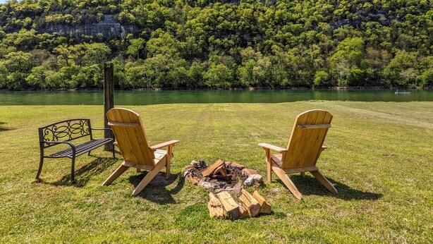 A grassy area with two wooden chairs and a bench facing a river, surrounded by dense green trees and a hill in the background. - The Lower Deck on Riverview ~ direct river access (Mountain View)