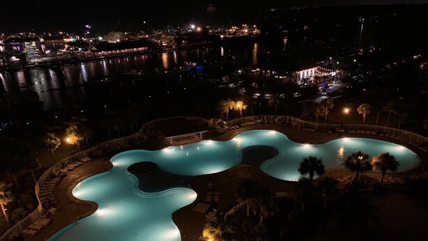 A nighttime view of a swimming pool complex with multiple interconnected pools, surrounded by lounge areas and palm trees.