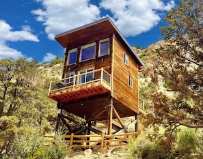 A wooden cabin with a balcony, built on stilts, surrounded by trees and rocky terrain.