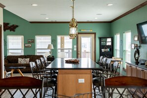 A dining area with a long table, chairs, and a chandelier. There is a sofa and a television in the background.