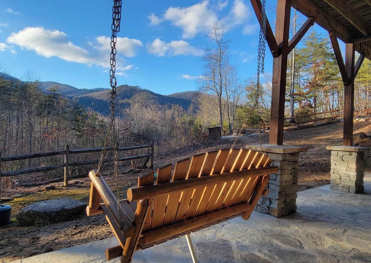 Un banc-balançoire en bois, placé sous une zone abritée et doté d’une base en pierre, avec en toile de fond un paysage montagneux.