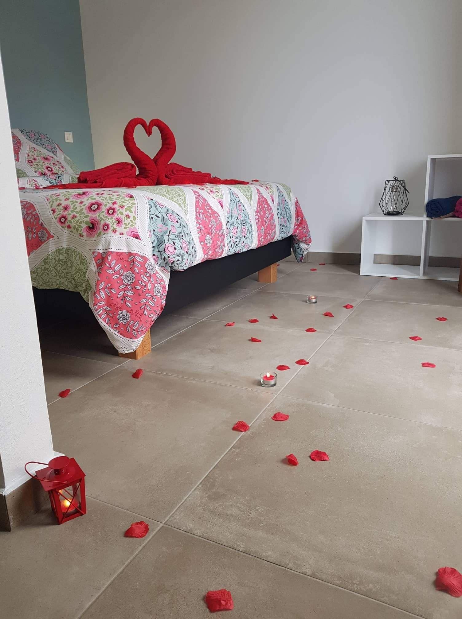Et soveværelse med en seng dækket af blomstret sengetøj, en hjerteformet pude og rosenblade spredt ud på gulvet.