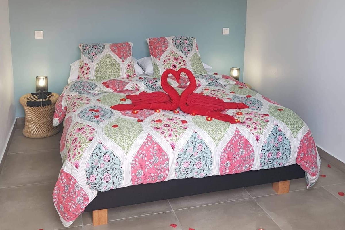 A bed with a floral quilt, red towels arranged in a heart shape, and rose petals scattered on the floor.