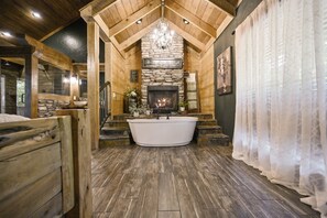 Une salle de bain rustique avec une cheminée en pierre, une baignoire sur pieds, un plancher en bois et un lustre.