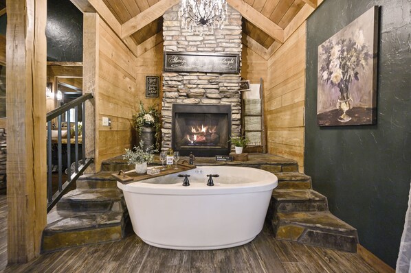Une cheminée en pierre avec un feu de bois, une baignoire blanche et un escalier menant à une porte en bois.