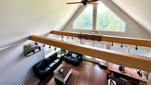 A loft-style room with a slanted ceiling, a ceiling fan, a wooden bench, a black leather sofa, a wooden coffee table, and a view of greenery through large windows.