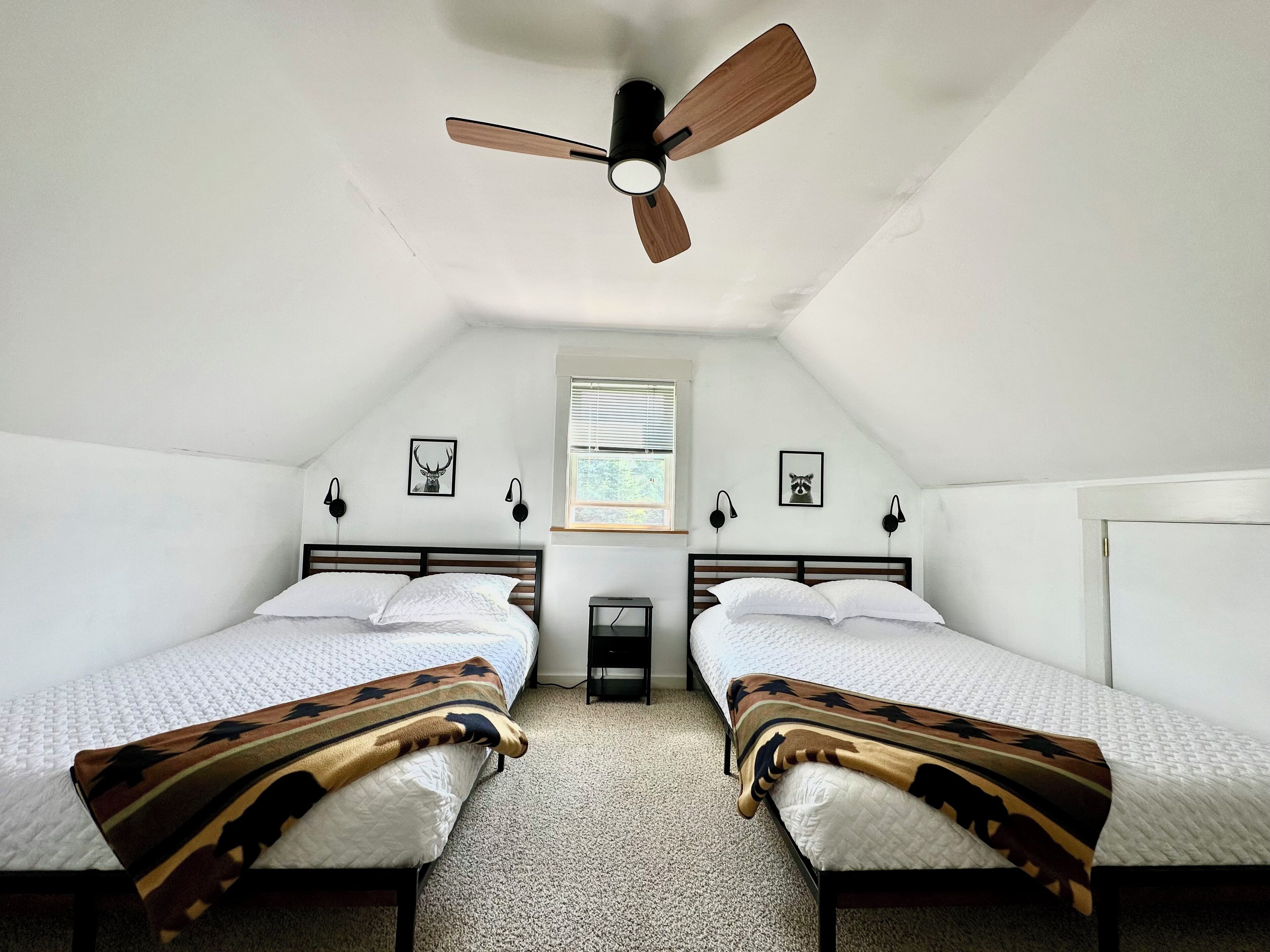 A room with two single beds, a ceiling fan, a window with blinds, and wall-mounted lamps.