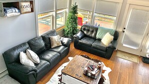 A living room with two black leather sofas, a wooden coffee table, and a cowhide rug. There is a Christmas tree and a view of a backyard through large windows.