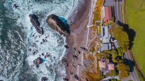 An aerial view of a coastal property with a beach, rocky shoreline, and a road leading to a cluster of buildings. - Seamist Beach Cottage (Bodega Bay)