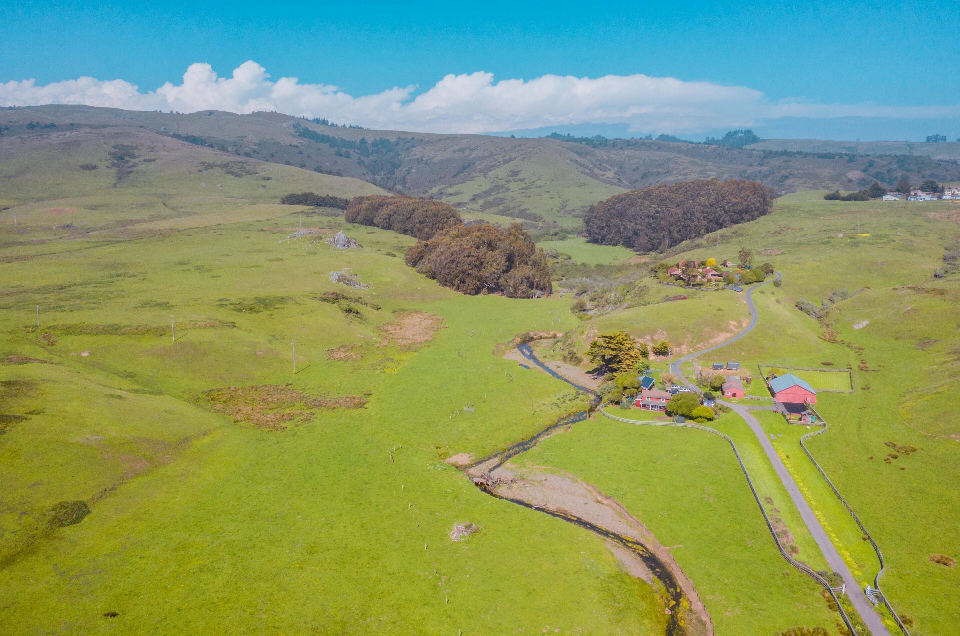 A rural landscape with rolling hills, a winding road, and a small cluster of buildings with red roofs.