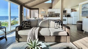 A spacious living area with a large window overlooking the ocean, a cozy seating area with a woven sofa, and a modern kitchen with white cabinets. - Seamist Beach Cottage (Bodega Bay)