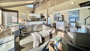 A spacious living area with a high ceiling, a dining area with a table and chairs, a kitchen with modern appliances, and a large window providing natural light. - Seamist Beach Cottage (Bodega Bay)