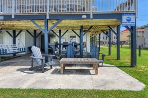 A covered outdoor seating area with Adirondack chairs and a wooden table, a rental sign, and a building with a balcony in the background.