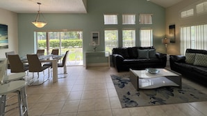 A spacious living area with a dining table, chairs, a sofa, and a coffee table. There are multiple windows providing natural light and a ceiling fan.