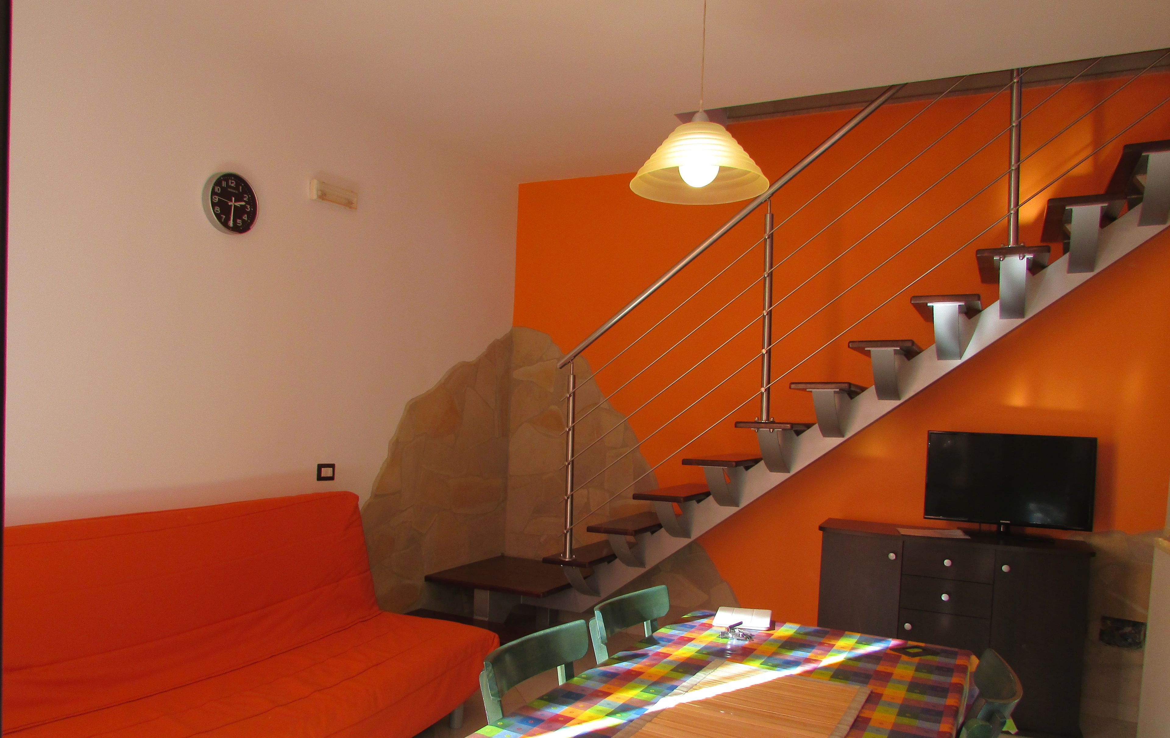 A modern living room with a red sofa, a colorful quilted table, a flat-screen TV, and a staircase with orange walls.