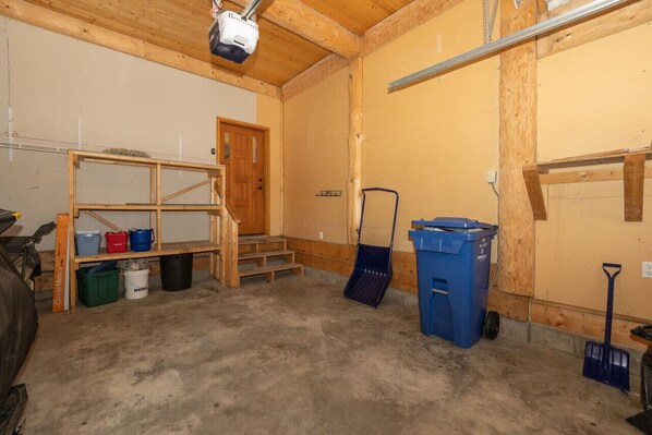 A room with a wooden ceiling, a door, a wooden shelf with buckets, a blue trash bin, a broom, and a concrete floor.