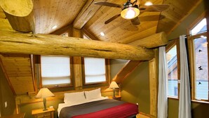 Une chambre avec un plafond en bois, un lit recouvert d’une couverture rouge, deux tables de chevet avec des lampes et des fenêtres avec des rideaux.