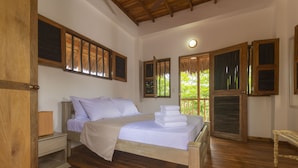A bedroom with a wooden bed, white linens, a nightstand, a wooden bench, and a window with shutters. - Samadhi Ecohotel by Rotamundos (Puerto Escondido)