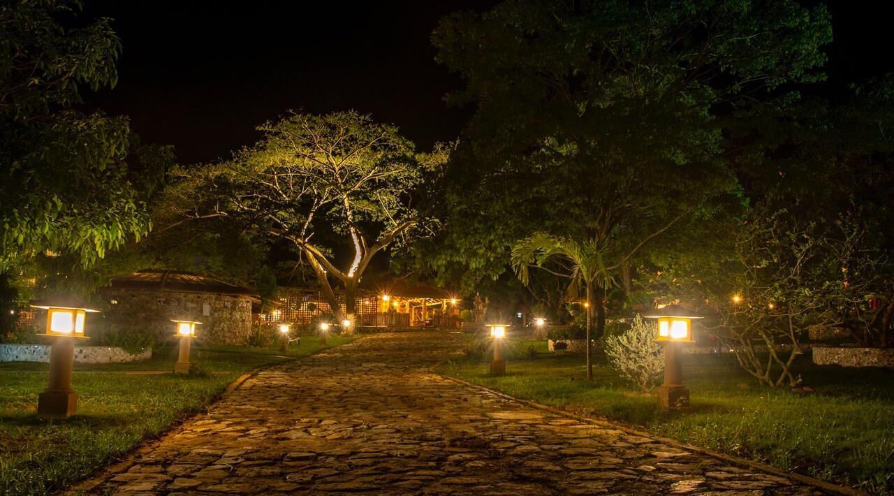 Un sendero de piedra conduce a un edificio flanqueado por faroles iluminados, rodeado de árboles y vegetación.