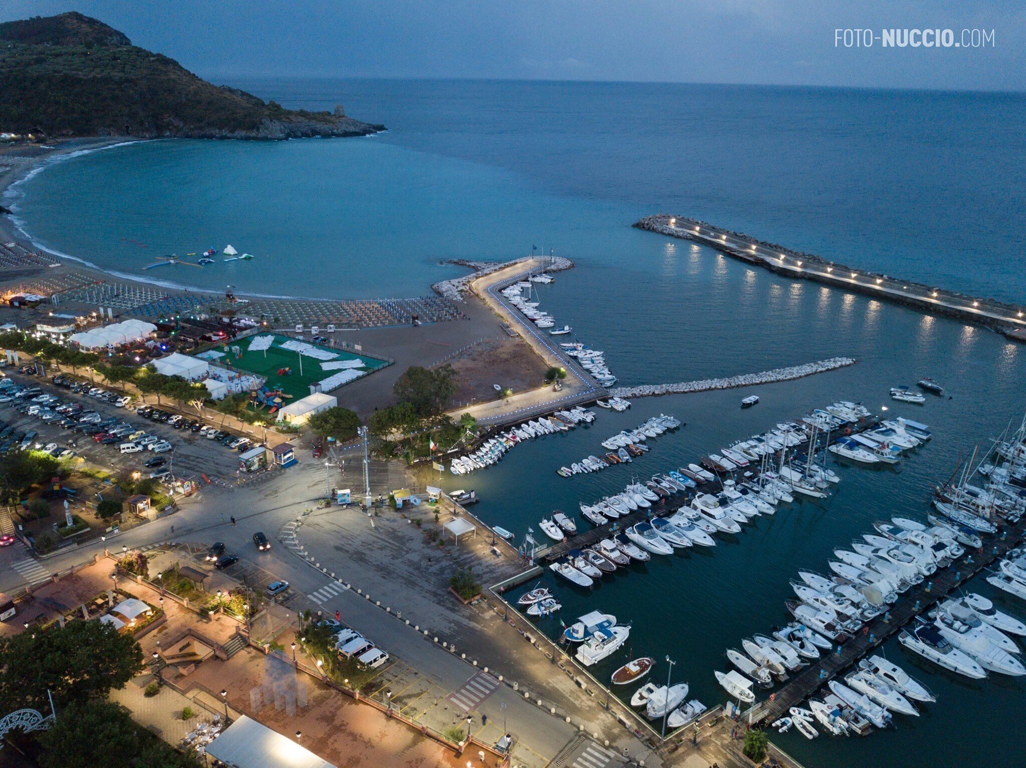 Vue aérienne d’une marina avec des bateaux amarrés, une route sinueuse et une zone de plage.