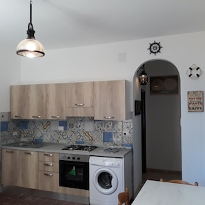 A compact kitchen with wooden cabinets, a gas stove, a washing machine, and a tiled backsplash. - Bellavista Central apartment with sea view  (Marina di Camerota SA)