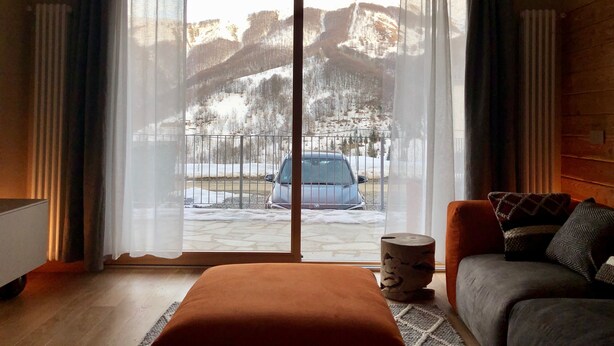 A hotel room with a large sliding glass door leading to a snowy outdoor area, a wooden floor, a bed with an orange headboard, and a sofa with patterned cushions. - 3 bedroom apartment Chalet Bizet (Panice Soprana)