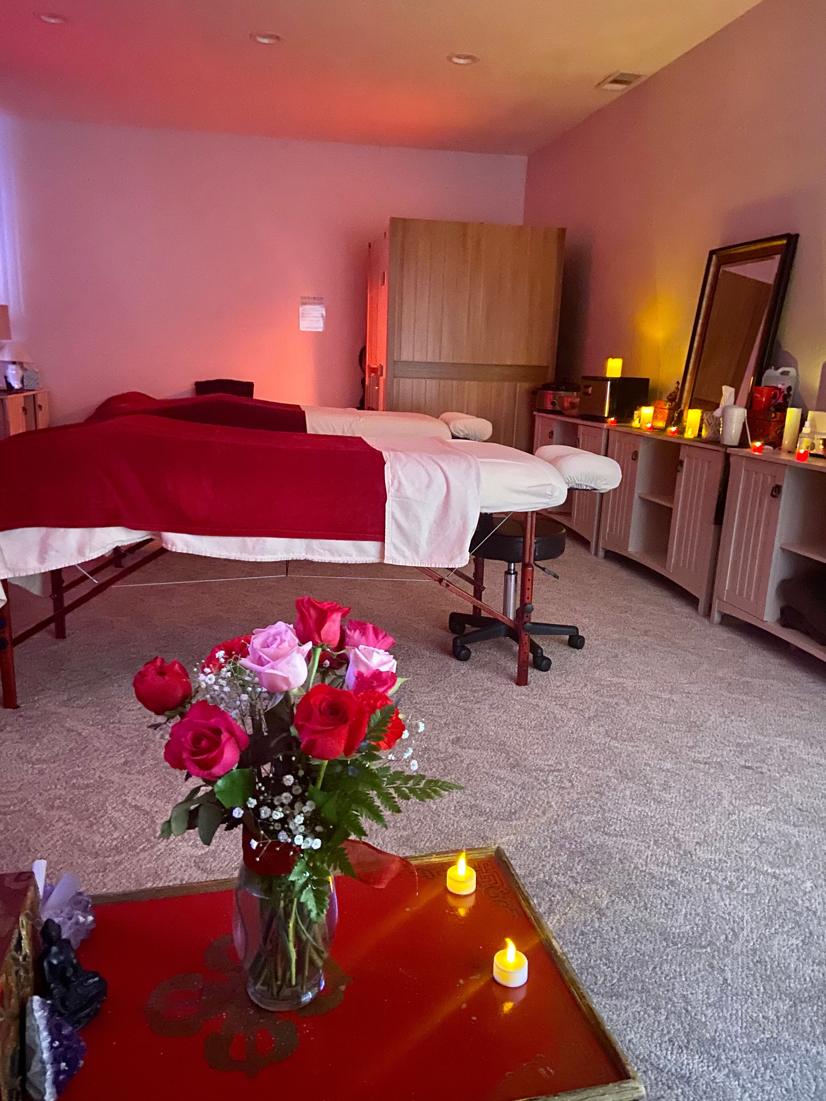 Una sala de masajes con dos camillas, una roja con un florero y velas.