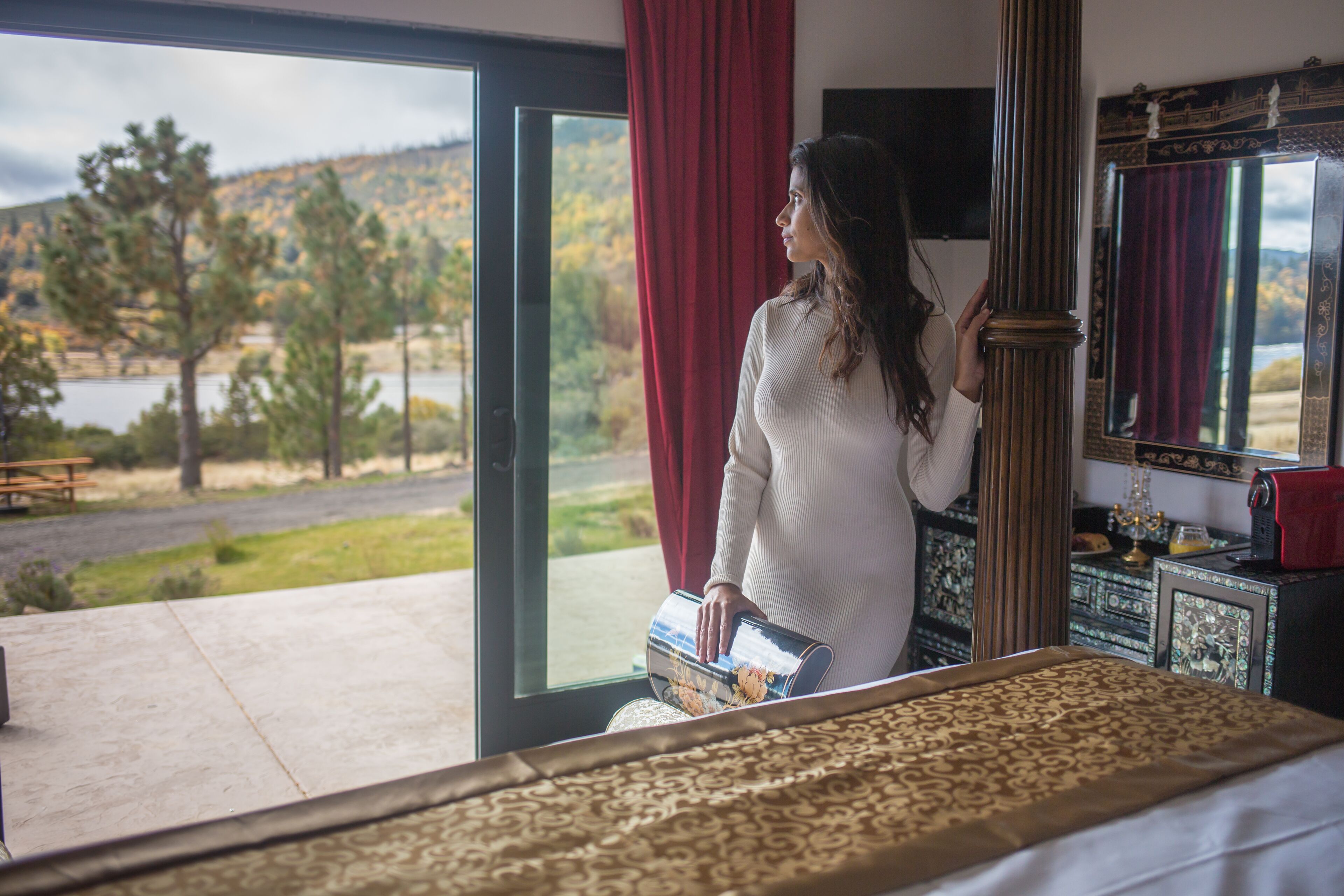 A room with a large window, a bed with a patterned bedspread, a wooden dresser, and a mirror with a decorative frame.